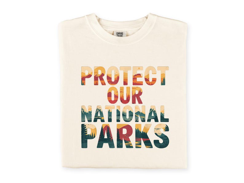 May include: A cream-coloured t-shirt with the words "PROTECT OUR NATIONAL PARKS" in a layered design. The text features a landscape of mountains, trees, and a sunset. The t-shirt is folded neatly.