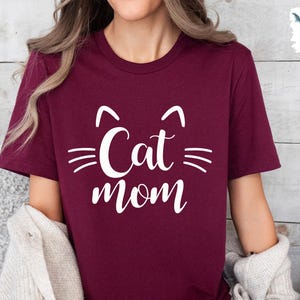 May include: A burgundy t-shirt featuring a white cat face design and the words "Cat Mom" in a script font. The design includes cat ears and whiskers. The shirt is made of a soft material.
