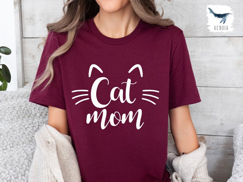 May include: A burgundy t-shirt featuring a white cat face design and the words "Cat Mom" in a script font. The design includes cat ears and whiskers. The shirt is made of a soft material.