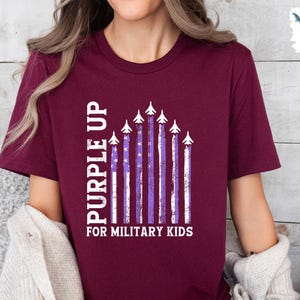 Op de afbeelding: Een maroon t-shirt met een witte en paarse afbeelding van een rij vliegtuigen die over een paarse en witte gestreepte Amerikaanse vlag vliegen. De tekst "Purple Up for Military Kids" staat onder de afbeelding.