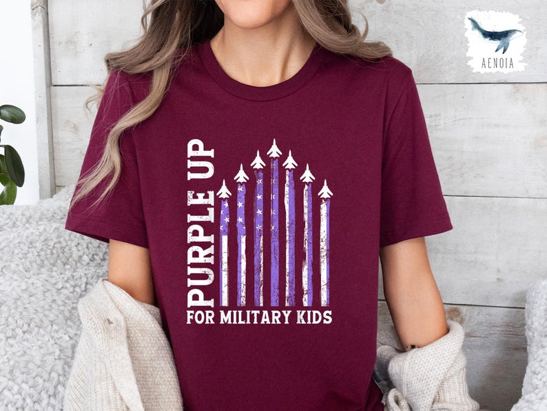 Op de afbeelding: Een maroon t-shirt met een witte en paarse afbeelding van een rij vliegtuigen die over een paarse en witte gestreepte Amerikaanse vlag vliegen. De tekst "Purple Up for Military Kids" staat onder de afbeelding.