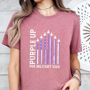 Op de afbeelding: Een mauve t-shirt met een witte afbeelding van een Purple Up-vlag met vliegtuigen en de tekst "Purple Up for Military Kids".