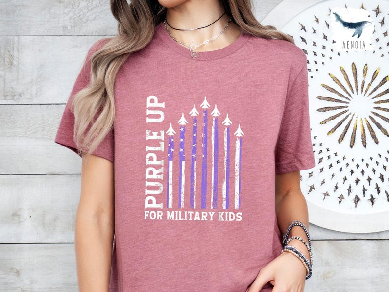 Op de afbeelding: Een mauve t-shirt met een witte afbeelding van een Purple Up-vlag met vliegtuigen en de tekst "Purple Up for Military Kids".