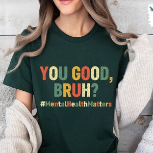 以下が含まれることがあります： 「YOU GOOD, BRUH?」とカラフルな文字で書かれた濃い緑色のTシャツ。メインテキストの下には「#MentalHealthMatters」というフレーズがあります。このシャツはカジュアルなスタイルです。
