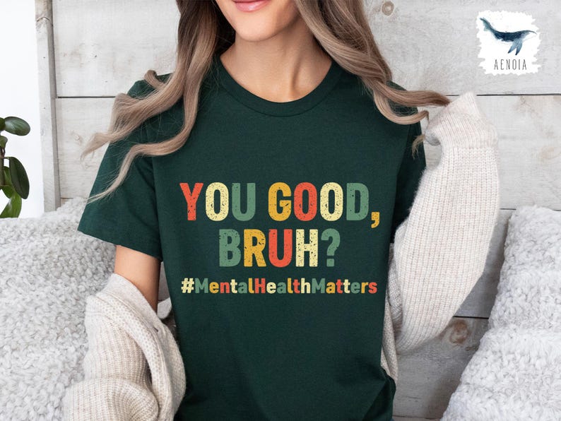 以下が含まれることがあります： 「YOU GOOD, BRUH?」とカラフルな文字で書かれた濃い緑色のTシャツ。メインテキストの下には「#MentalHealthMatters」というフレーズがあります。このシャツはカジュアルなスタイルです。