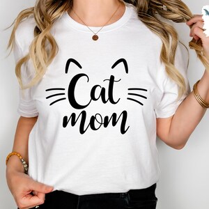 May include: White t-shirt featuring the text "Cat mom" in black, with cat ear and whisker graphics. A casual, everyday wear t-shirt. The design is simple and bold.