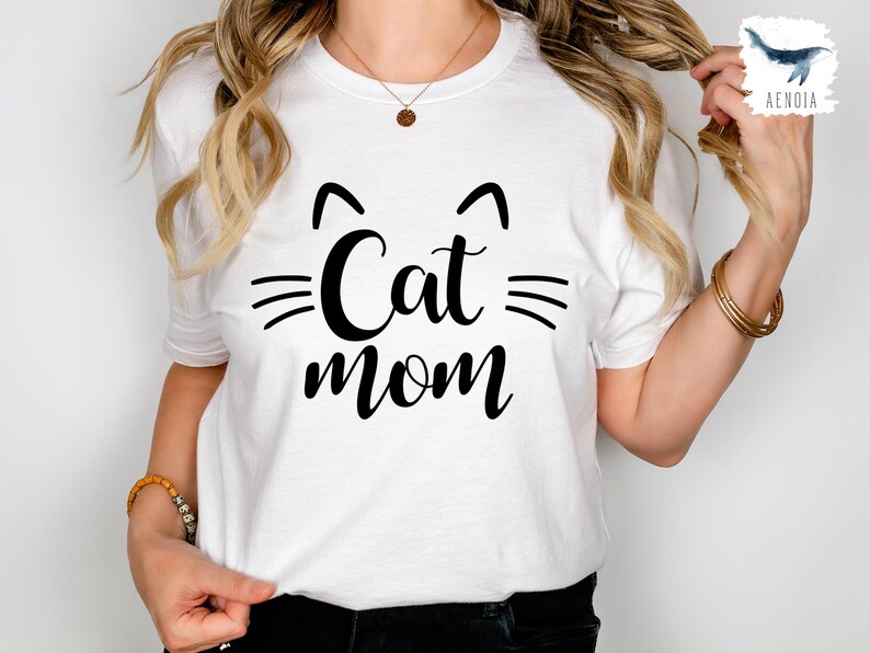 May include: White t-shirt featuring the text "Cat mom" in black, with cat ear and whisker graphics. A casual, everyday wear t-shirt. The design is simple and bold.