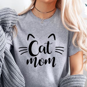 May include: A grey t-shirt featuring the text "Cat Mom" in a black, stylised font. The design includes cat ears above the word "Cat" and whiskers. The shirt is made of a soft material.