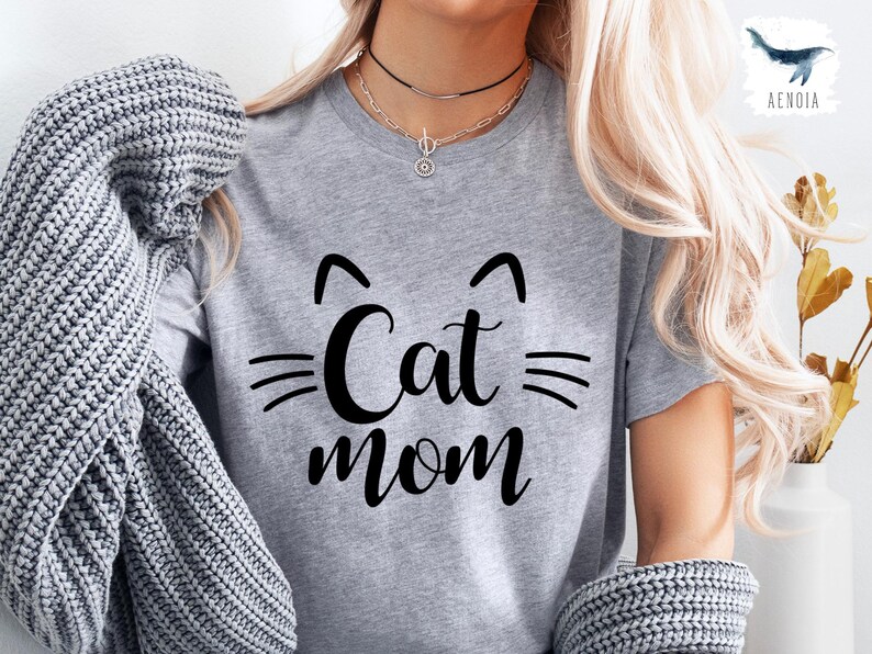 May include: A grey t-shirt featuring the text "Cat Mom" in a black, stylised font. The design includes cat ears above the word "Cat" and whiskers. The shirt is made of a soft material.
