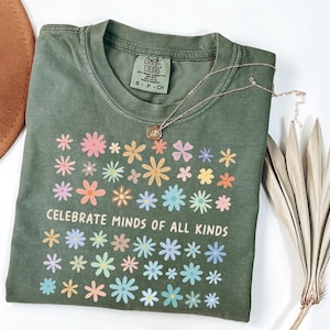 May include: An olive green t-shirt with a floral design and the text "CELEBRATE MINDS OF ALL KINDS". The shirt has a crew neck and short sleeves. A gold necklace with a pendant is on the shirt.