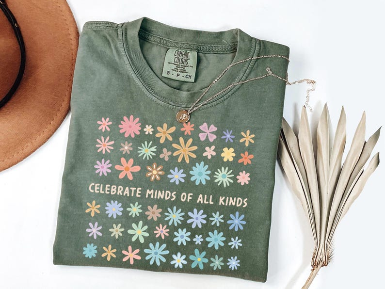 May include: An olive green t-shirt with a floral design and the text "CELEBRATE MINDS OF ALL KINDS". The shirt has a crew neck and short sleeves. A gold necklace with a pendant is on the shirt.