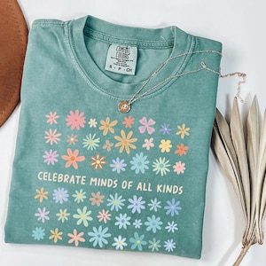 May include: A teal Comfort Colors t-shirt with a floral design and the text "CELEBRATE MINDS OF ALL KINDS." A silver necklace with a pendant is on the shirt. A brown hat and dried plant are in the background.