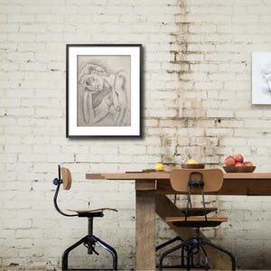 May include: A black framed drawing of a person lying down with their head resting on their arm. The drawing is on a white background and is hanging on a white brick wall.