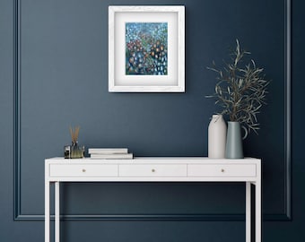 May include: A framed abstract art print in a white wooden frame. The artwork has a colourful, textured design with blues, greens, and reds. The frame is displayed on a white console table against a dark blue wall.