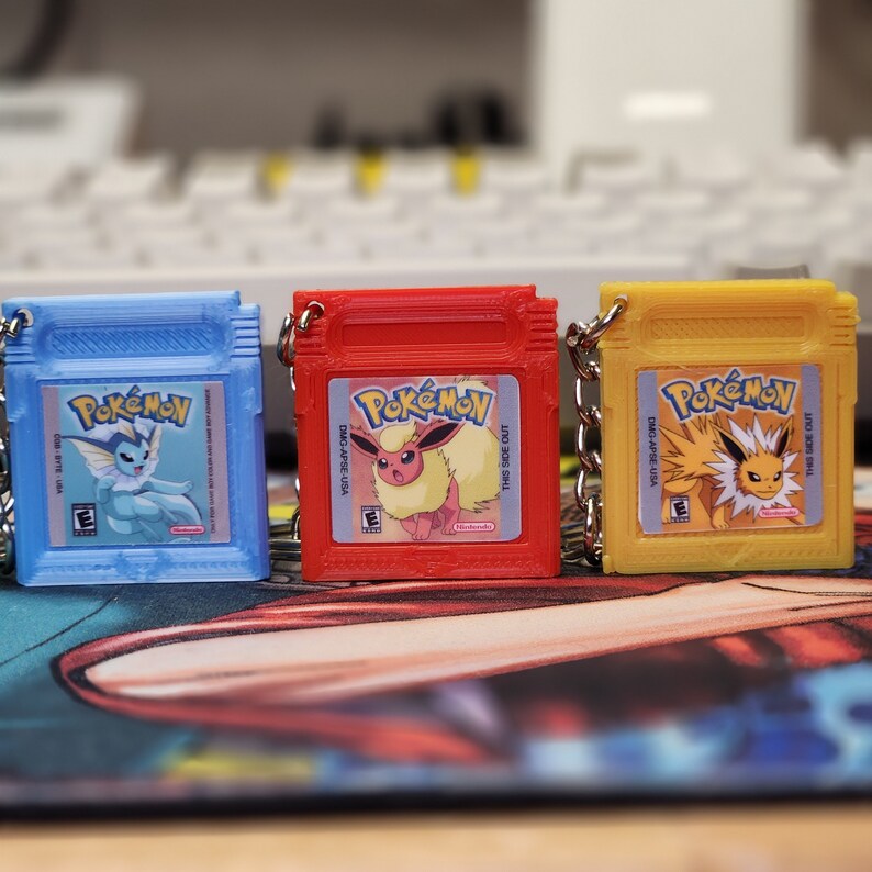 May include: Three Pokemon game cartridge keychains. The keychains are blue, red, and yellow. Each keychain features a different Pokemon character. The text on the keychains reads "Pokemon" and "This Side Out".