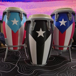 May include: Three conga drums with the Puerto Rican flag design. The drums are painted in red, white, and blue, with a white star. The drums are on gray stands.