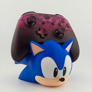 May include: A blue Sonic the Hedgehog-themed controller holder with a black and pink controller resting on top. The holder features Sonic's face and ears, with a white belly and tan muzzle. The controller has black buttons and a pink gradient.