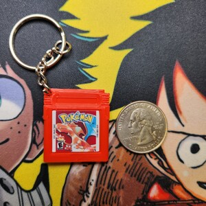 May include: A red Game Boy Color keychain with a Pokemon game cartridge design. The keychain features the text "Pokemon" and a picture of a Charizard.
