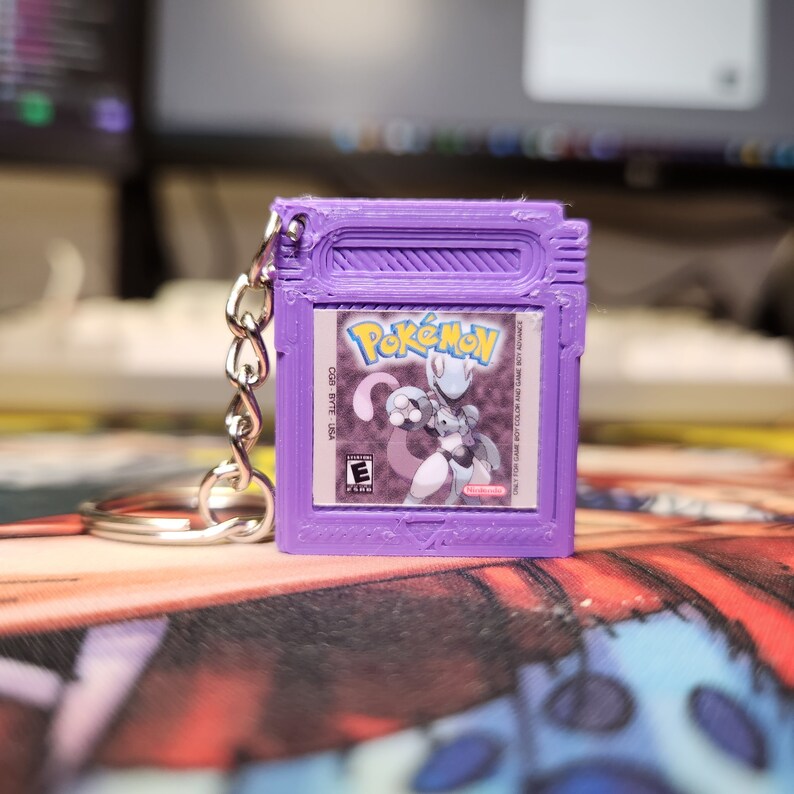 May include: Purple Pokemon Game Boy Color keychain with a picture of Mewtwo on the front. The keychain has a silver metal ring.