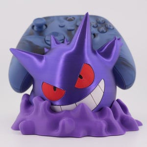 May include: A purple Gengar-themed controller stand with red eyes and a wide grin. The stand is holding a blue and black marbled Xbox controller. The stand has a wavy base and is designed to hold a gaming controller.