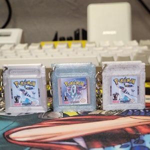 May include: Three silver Pokemon Game Boy Color game cartridges with keychains. The cartridges are decorated with glitter and feature the Pokemon logo and a Pokemon character on the front. The text on the cartridges reads "Pokemon" and "Game Boy Color".