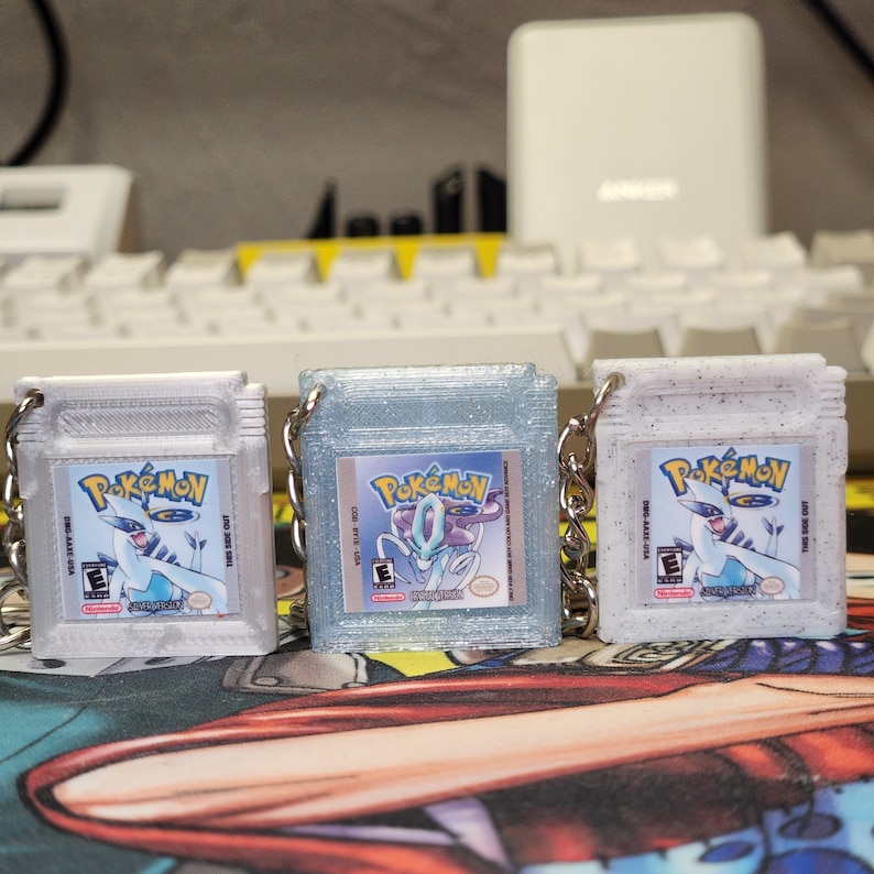 May include: Three silver Pokemon Game Boy Color game cartridges with keychains. The cartridges are decorated with glitter and feature the Pokemon logo and a Pokemon character on the front. The text on the cartridges reads "Pokemon" and "Game Boy Color".