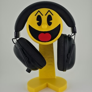 May include: A yellow Pac-Man-themed headphone stand with black headphones. The stand features a smiling Pac-Man face with a red mouth and black eyes. The headphones are black with padded earcups.