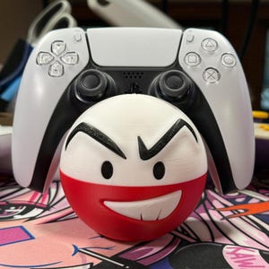 May include: A white and black PlayStation 5 controller with a red and white 3D printed Poké Ball character with a frowning face.