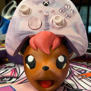 May include: A pink and purple swirl Xbox controller with white buttons is sitting on top of a brown 3D printed Pokemon character head.