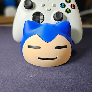 May include: A white Xbox controller with black joysticks and colored buttons is positioned behind a blue and tan Snorlax figure. The Snorlax figure has a smiling face and is resting on a dark blue surface.