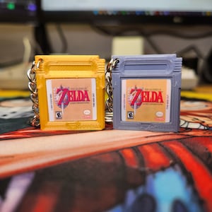 May include: Two yellow and gray keychains shaped like Game Boy cartridges. The keychains feature the Nintendo game "The Legend of Zelda: Link's Awakening".