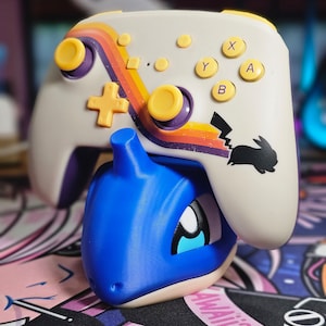 May include: A white and yellow Nintendo Switch controller with a Pikachu design on the back. The controller is sitting on top of a blue and white Pokemon figurine.