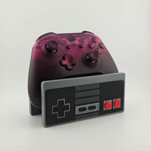 May include: A black and pink video game controller with a retro-style gray controller stand. The controller has a pink and black gradient design. The stand has a classic gray, black, and red color scheme.