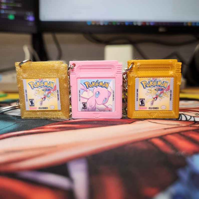 May include: Three gold, pink, and yellow Pokemon Game Boy cartridge keychains. Each keychain features a different Pokemon character on the front. The keychains are made of plastic and have a metal ring for attaching to keys.