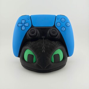 May include: A blue PlayStation 5 controller rests on a black dragon-themed stand with green eyes. The controller has blue buttons and a black center. The dragon stand has a detailed, textured design.