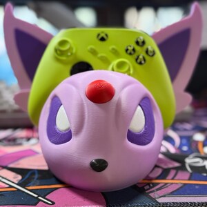 May include: A green and purple 3D printed controller stand shaped like a Pokemon character. The stand is designed to hold a gaming controller.