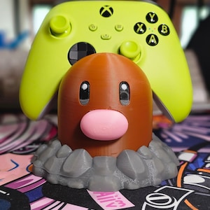 May include: A green Xbox controller is held in place by a brown and gray 3D printed stand shaped like a Pokemon character. The stand has a pink nose and black eyes.