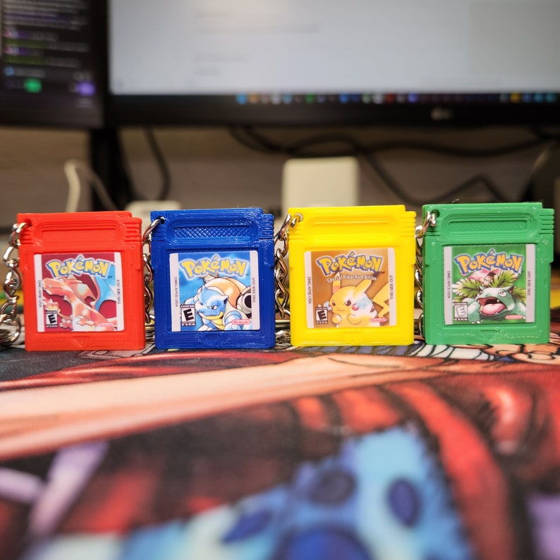 May include: Four different colored keychains featuring the Pokemon logo and characters. The keychains are shaped like the original Game Boy cartridges. The keychains are red, blue, yellow, and green.