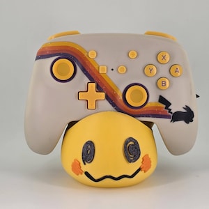 May include: A custom-designed video game controller with a retro rainbow stripe design and yellow accents. The controller sits atop a yellow Mimikyu-shaped stand. The controller has a black rabbit silhouette and the letters X, Y, A, and B.