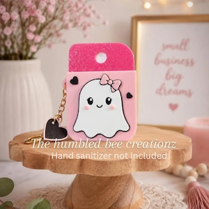 May include: A light pink hand sanitizer holder featuring a white ghost character with a small pink bow, black eyes, and rosy cheeks. The holder has a glittery pink top section, two small black heart accents, and a gold chain with a black heart charm.