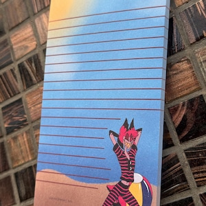 May include: A notepad with a beach scene illustration. The notepad features a blue sky with horizontal lines, a sandy beach, and a cartoon character in red and black stripes leaning on a beach ball.