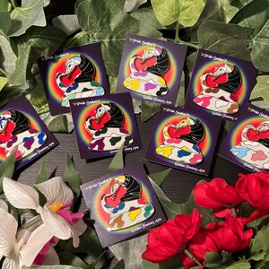 May include: Eight enamel pins featuring a pride snake design. Each pin features a different colour scheme with a rainbow background. The pins are all on a dark surface with flowers.