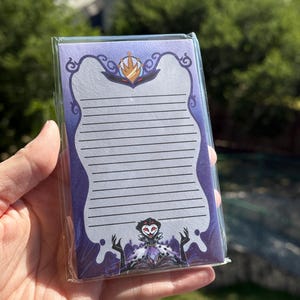 May include: A lined notepad with a purple border and a decorative top and bottom design. The notepad features a character illustration at the bottom. The notepad is in a clear plastic wrapper.