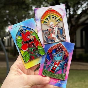 May include: Three enamel pins with stained glass designs. One pin shows a red character with roses, another a figure with a rainbow and roses, and the third a blue character. Each pin is on a colourful card with the text "Ace of Roses" and "Golden Fantasy Art".