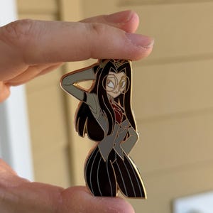 May include: A gold-coloured enamel pin depicting a stylized female character with long black hair, wearing a grey suit jacket, a red top, and black and white striped trousers. The character is posed with one hand on her head.