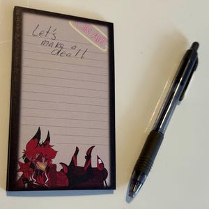 May include: A black notepad with a red and black cartoon character on the bottom. The notepad has lined paper and the words "Let's make a deal!" written on it. There is a pink sticker on the top right corner that says "ON AIR".