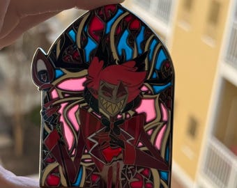 May include: A stained-glass style enamel pin featuring a character with red hair and a red suit. The pin has a black outline and is set against a pink, blue, and red background. The pin is held up by a hand.