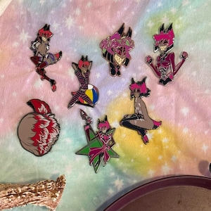 May include: A collection of seven enamel pins featuring cartoon characters in various poses. The pins showcase a color palette of pink, red, black, and white. One pin depicts a stylized wolf, while others feature characters with horns and sharp teeth.