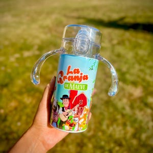 Puede incluir: Un vaso para beber transparente y azul con asas. El vaso presenta una colorida ilustraci&oacute;n de una escena de granja con el texto "La Granja de Maeve". El dise&ntilde;o incluye personajes de dibujos animados, un granero rojo y un vaquero tocando la guitarra.