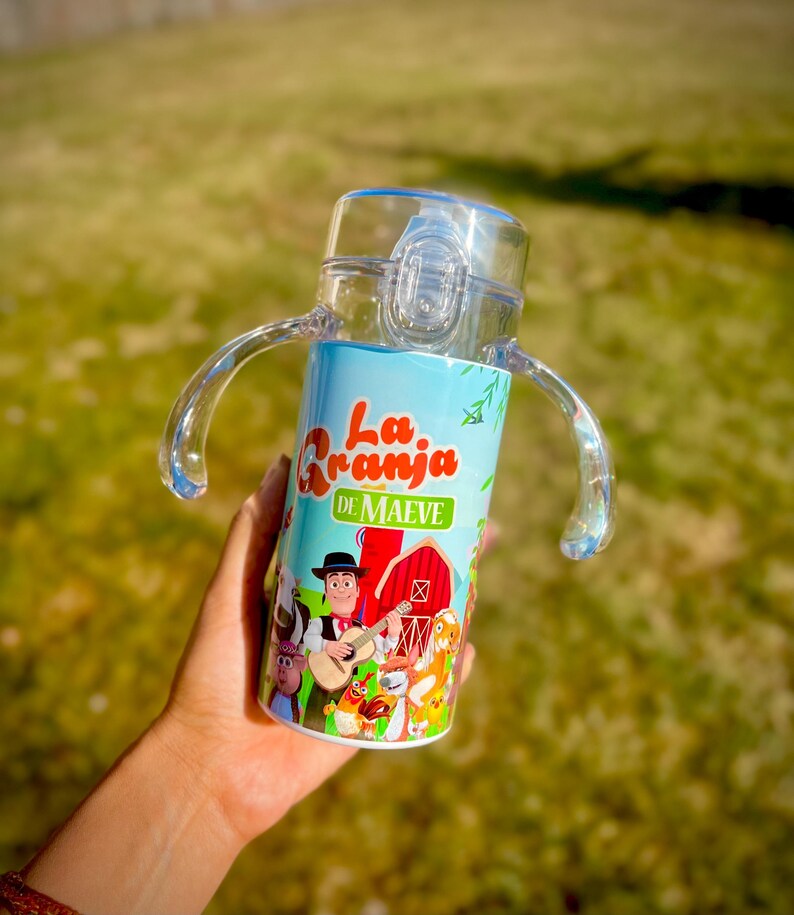 Puede incluir: Un vaso para beber transparente y azul con asas. El vaso presenta una colorida ilustraci&oacute;n de una escena de granja con el texto "La Granja de Maeve". El dise&ntilde;o incluye personajes de dibujos animados, un granero rojo y un vaquero tocando la guitarra.